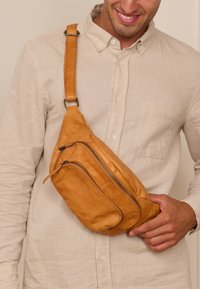 Lederumhängetasche in Tan mit einer geschwungenen Form. Verfügt über zwei Reißverschlüsse und einen verstellbaren Trageriemen. Wird über den Körper getragen. Glatte Textur.