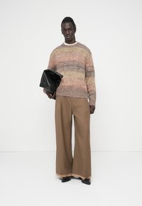 Πλεκτό πουλόβερ σε πολυχρωμίες παστέλ αποχρώσεις, άνετη εφαρμογή. Φαρδιά καφέ παντελόνια με ελαφριά γυαλάδα, μαύρη τσάντα στο χέρι, γυαλιστερά παπούτσια.