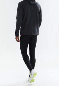 Hoodie de sport noir à manches longues avec un détail zipper dans le dos, associé à un legging noir ajusté et des baskets blanches à semelles vertes.