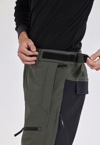 Verstelbare cargobroek van donkergroen nylonmateriaal, met een zwart zijpaneel, klittenbandsluiting en meerdere zakken.