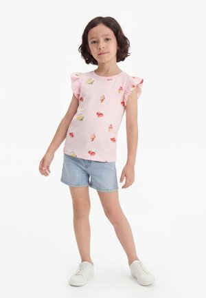 Ung pige med kort mørkt hår iført en lyserød top med flæser og isprint, lyse denimshorts og hvide sneakers, stående.