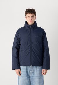 Marine pufferjack die is voorzien van een hoge kraag, een ritssluiting aan de voorkant en zijzakken. Het materiaal heeft een gladde textuur met een subtiele glans.