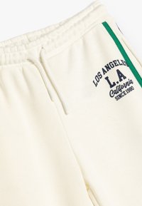 Pantalones de chándal color crema con cordón, que presentan una franja lateral verde y el texto "Los Ángeles L.A. California Desde 1980" en azul marino.