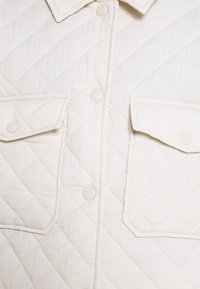 Kremowa pikowana kurtka z dwoma kieszeniami na piersi, zatrzaskami i usztywnianym kołnierzem. Tkanina ma miękką, teksturowaną powierzchnię.