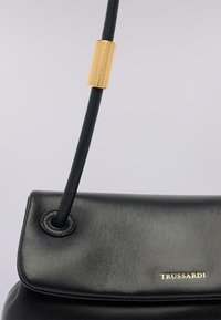 Černá kožená kabelka s přehnutým klopou, zlatými kovovými doplňky s názvem značky a elegantním, minimalistickým designem.