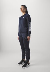 Námořnická zipová mikina a odpovídající tepláky s bílým logem Adidas na rukávu a malým logem na hrudi, doplněné černými sportovními botami.