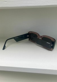 Lunettes de soleil avec une monture sombre, des verres transparents roses, une forme angulaire et un design épuré, arborant la marque "KOMONO" sur le côté.