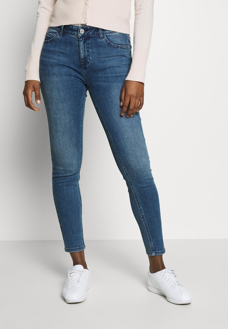 Femme portant un jean skinny bleu, des baskets blanches et un cardigan beige clair, debout devant un fond clair uni.