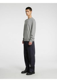 Grå strikkegenser med lange ermer og rund hals, kombinert med løse mørke bukser og svarte sko, stående foran en ensfarget bakgrunn.