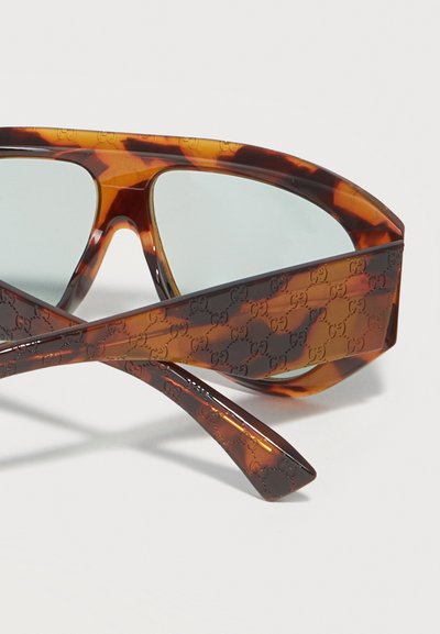 Γυαλιά ηλίου με μοτίβο χελώνας, με χοντρά πλαίσια και φακούς ανοιχτού μπλε χρώματος, που φέρουν επαναλαμβανόμενο ανάγλυφο λογότυπο σχεδιαστή στα πλαϊνά μέρη.