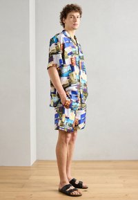 Kortærmet, knap-op skjorte og matchende shorts lavet af stof med farverige postkortmotiver. Sorte sandaler fuldender outfittet.