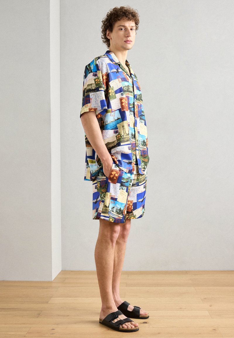 Kortærmet, knap-op skjorte og matchende shorts lavet af stof med farverige postkortmotiver. Sorte sandaler fuldender outfittet.