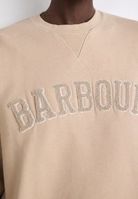 Beige sweatshirt featuring textured, embroidered "BARBOUR" lettering in a contrasting color. Round neckline with cross-stitch detail at shoulder.