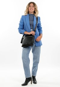 Frau trägt ein blaues Sakko, graues Oberteil, hellblaue Jeans, schwarze Stiefel mit Absatz und eine schwarze Umhängetasche, lächelt und steht.