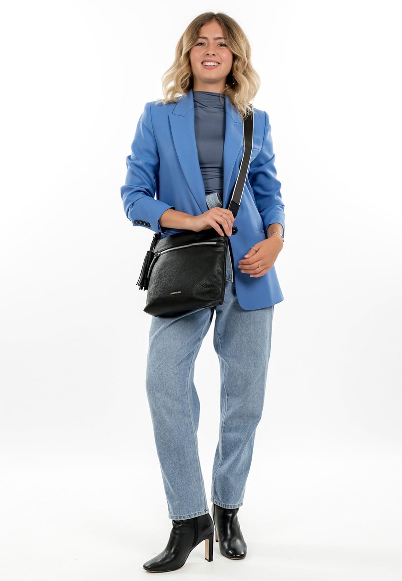 Frau trägt ein blaues Sakko, graues Oberteil, hellblaue Jeans, schwarze Stiefel mit Absatz und eine schwarze Umhängetasche, lächelt und steht.