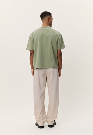 Μπλούζα από βαμβάκι σε ελιά πράσινο χρώμα με κοντά μανίκια, φαρδιά κοπή και χωρίς εμφανή λογότυπα, συνδυασμένη με φαρδιά παντελόνια σε κρεμ χρώμα και μαύρα παπούτσια.