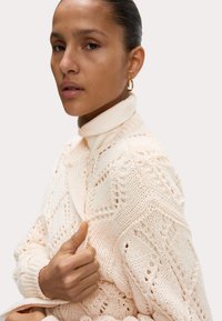 Strickpullover in hellem Creme mit geometrischen Mustern, geripptem Kragen und Bündchen; Modell hält das Pulloverstück nahe am Ausschnitt.