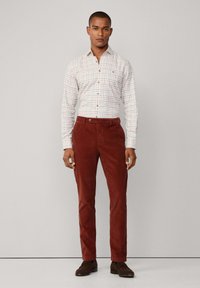 Geruit shirt in wit, rood en blauw; bruine ribfluwelen broek; staande mannelijke figuur; bruine schoenen; neutrale achtergrond.