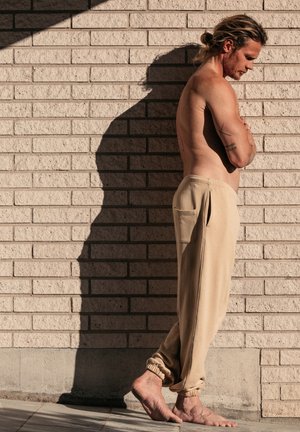 Beige sweatpants met elastische boorden en zijzakken, staand tegen een gestructureerde bakstenen muur, waarbij een relaxte pasvorm en gladde stof worden getoond.