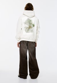 Crèmekleurige hoodie met een groot groen abstract ontwerp op de achterkant, gecombineerd met bruine ribfluwelen broek. Soepele stof met een relaxte pasvorm.