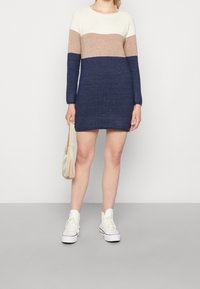 Robe en maille avec un col rond, présentant un bloc de couleurs en crème, beige et marine. Manches longues, longueur au-dessus du genou, portée avec des baskets blanches.