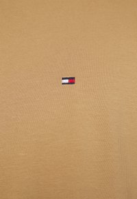 T-shirt en coton beige avec un tissu texturé et un petit logo brodé aux accents rouge, blanc et bleu sur le côté gauche de la poitrine.