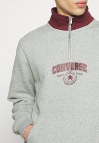 Szara bluza z marnowym półzamkiem. Wyróżnia się napisem "CONVERSE" dużymi literami oraz nadrukiem logo. Wykonana z miękkiego materiału, z kieszenią typu kangur.