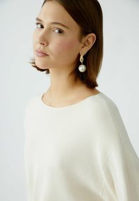 Weißes Rippenshirt mit weitem Ausschnitt, kombiniert mit großen Ohrringen in goldfarbener Perle. Weiche Textur und lockere Passform.