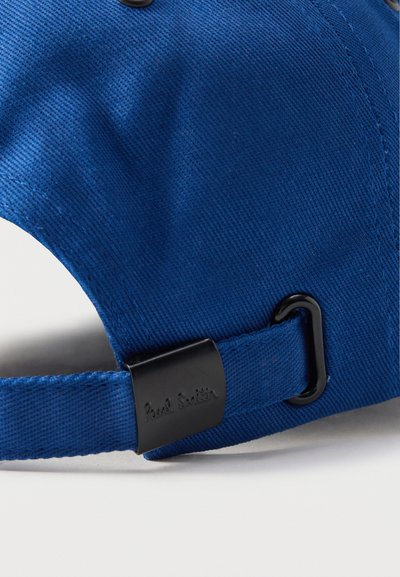 Μπλε καπέλο με υφασμάτινη υφή, ρυθμιζόμενο λουράκι και μεταλλικά εξαρτήματα με χαραγμένο λογότυπο στο λουράκι.