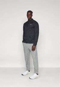 Zwarte Nike-jas met een hoge kraag, grijze joggingbroek en witte schoenen met oranje accenten. Soepele stof, sportieve pasvorm, minimalistisch ontwerp.