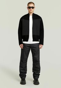 Giacca nera con zip e dettaglio a pannelli, indossata sopra una maglietta bianca. Abbinata a pantaloni di jeans scuri e scarpe nere. Il modello è in piedi.