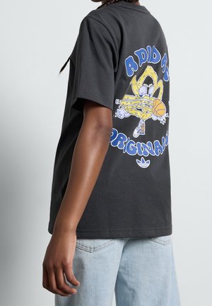 Person trägt ein schwarzes Adidas Originals T-Shirt mit einem Cartoon-Maskottchen, das einen Basketball dribbelt, kombiniert mit hellblauen Jeans.