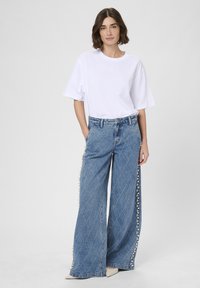 Žena v voľnom bielom tričku a širokých džínsoch s diamantovým prešívaním a perlovými detailmi pozdĺž strán.