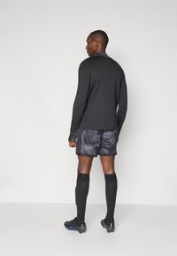 Langermet svart treningsskjorte kombinert med grå kamuflasje-shorts, svarte knelengde sokker og svarte fotballsko med blå detaljer.