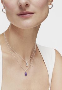 Collares de capas de plata con charms distintos: un pequeño colgante de plata y un colgante de gema púrpura. Textura suave, cadenas delicadas.