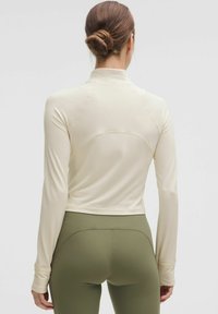 Tricou cu maneci lungi în crem deschis, cu un model texturat în zigzag, având un guler înalt și un design mulat, asortat cu colanți verzi măslinii.