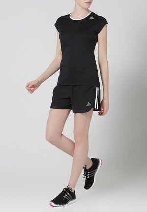 Femme portant une tenue sportive Adidas noire composée d'un short, d'un haut à manches courtes ajusté, et de chaussures de course noires avec des semelles blanches et roses.