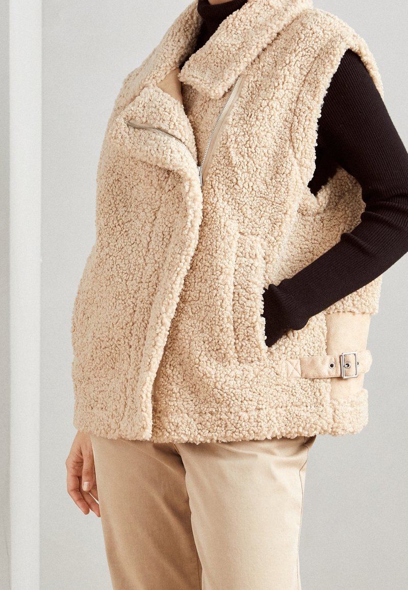 Veste sans manches en polaire sherpa beige avec fermeture éclair asymétrique, boucle latérale, portée sur un haut noir à manches longues et un pantalon beige, main dans la poche.