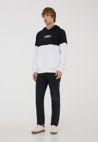 Felpa con cappuccio, parte superiore nera e parte inferiore bianca, materiale in fleece, tasca frontale e logo stampato. Abbinata a pantaloni scuri e sneakers bianche.