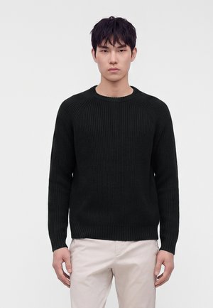 Jeune homme portant un pull en maille côtelée noire et un pantalon beige clair, debout devant un fond gris clair uni.