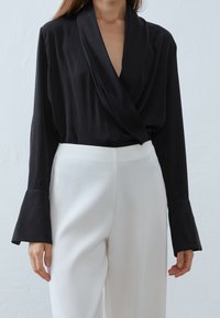 Top nero a maniche lunghe con scollatura drappeggiata e polsini a campana, abbinato a pantaloni bianchi a vita alta. Texture del tessuto liscia.
