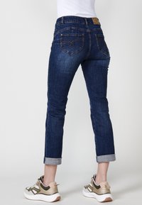 Temno modri traper hlače z oprijetim krojem in zavihanimi manšetami, s dekorativnim šivanjem na zadnjih žepih ter rjavim usnjenim označevalnikom v pasu.