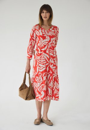 Kobieta w czerwono-białej kwiecistej sukience midi, trzyma brązową zamszową torebkę i nosi beżowe płaskie buty na jednolitym tle.