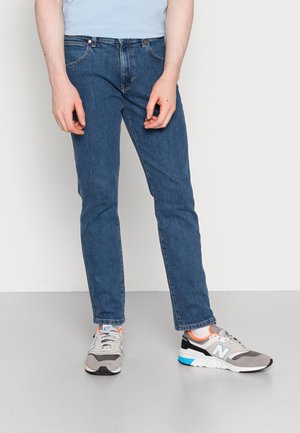 Person iført blå jeans og grå New Balance sneakers står mod en ensfarvet hvid baggrund med hænder afslappet ned langs siderne.