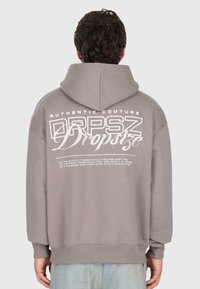 Muž oblečený v šedej mikine s kapucňou, na ktorej je na chrbte vytlačený nápis "Dropsize" a motivačný text, stojí proti jednoduchému šedému pozadiu.