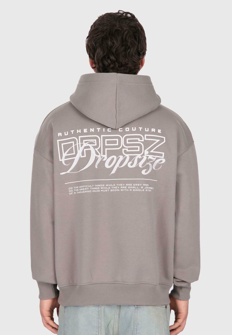 Muž oblečený v šedej mikine s kapucňou, na ktorej je na chrbte vytlačený nápis "Dropsize" a motivačný text, stojí proti jednoduchému šedému pozadiu.