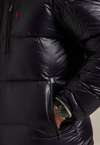 Black shiny puffer jacket with red logo, partially unzipped pocket showing wrist wearing green-faced watch with black strap.