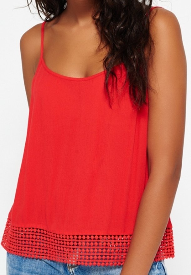 Czerwony top bez rękawów wykonany z lekkiego materiału, z delikatnym, teksturowanym wykończeniem na szydełku w kształcie trójkątnych wzorów, zestawiony z krótkimi szortami jeansowymi.