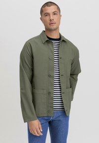 Olijfgroen lichtgewicht jack met een knoopsluiting, twee voorzakken en een kraag, gedragen over een marineblauw en wit gestreept shirt.