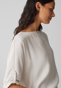 Hellbeige, lockere Bluse mit einem Bootsausschnitt, Dreiviertelärmeln und umgeschlagenen Bündchen, gefertigt aus einem strukturierten Stoff.